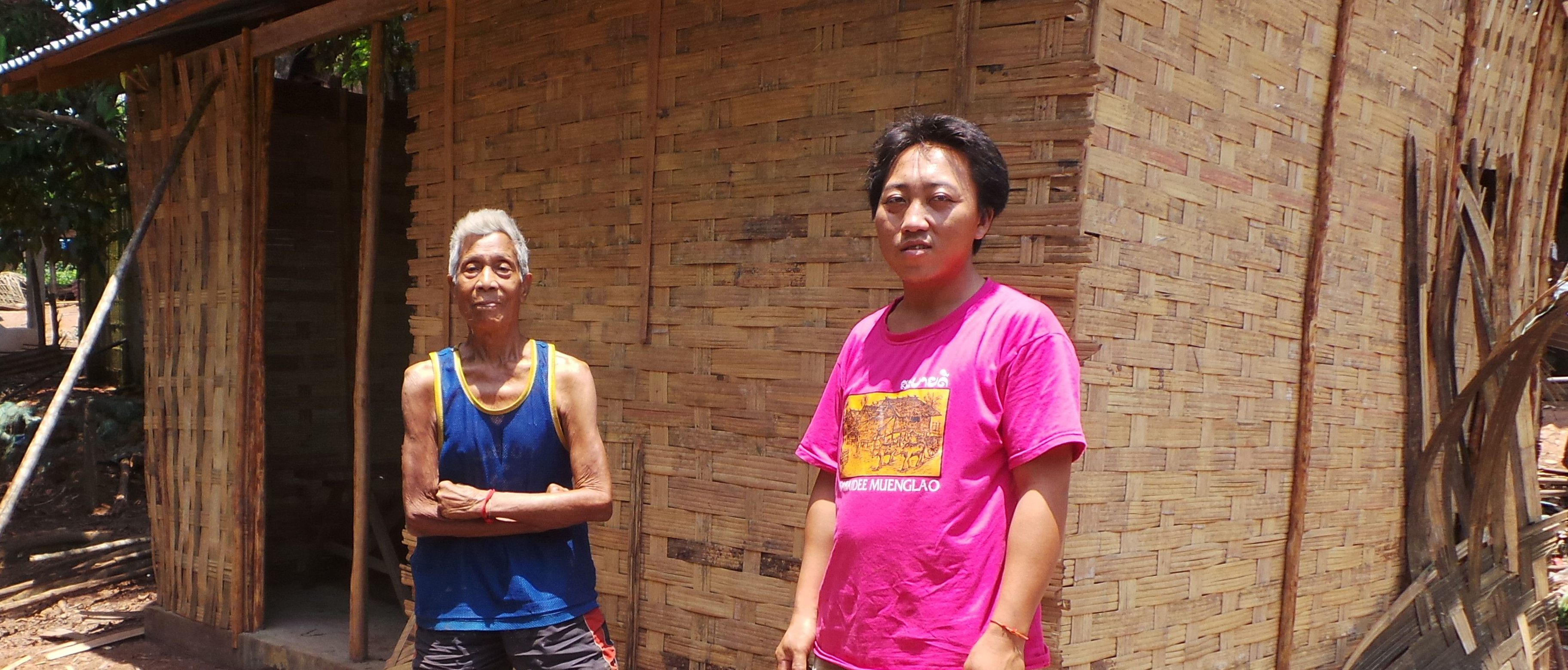 Eine helle geflochtene Wand aus Bambusblättern ziert das neue Haus. Herr Kham steht mit einem roten Hemd vor dem Haus. Seine Frau steht im Hintergrund.