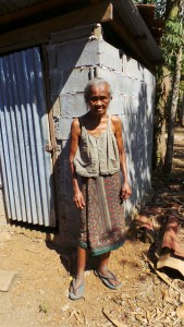 Eine Frau steht vor einer mit grauen Steinen gemauerte Toilette, welche mit einer Wellblechtür verschlossen ist.