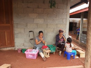Zwei Frauen sitzen vor einem gemauerten Haus und beschäftigen sich mit Handarbeit. Ein junges Mädchen schaut zu.